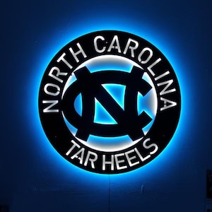 May include: A circular, black metal sign with the words "NORTH CAROLINA" and "TAR HEELS" encircling a blue and white logo. The sign is illuminated with a blue LED light, mounted on a dark blue wall. The image shows a gaming setup with two monitors.