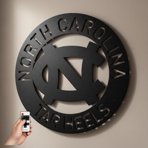 May include: A black metal wall art piece featuring the words "NORTH CAROLINA" and "TAR HEELS" cut out around a central logo. The logo is a stylized "NC" design. A hand holds a remote control in the lower left corner.