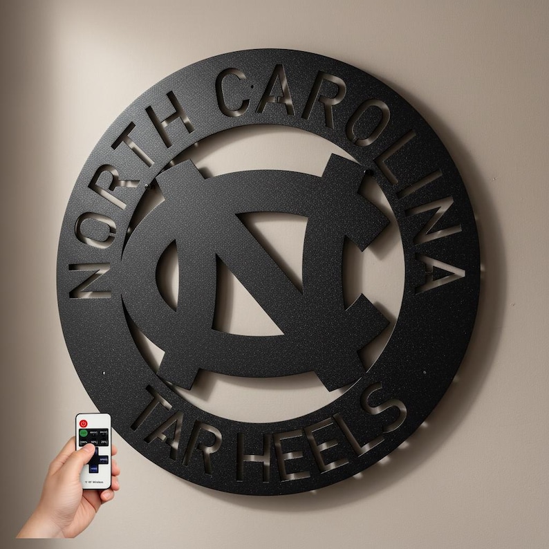 May include: A black metal wall art piece featuring the words "NORTH CAROLINA" and "TAR HEELS" cut out around a central logo. The logo is a stylized "NC" design. A hand holds a remote control in the lower left corner.