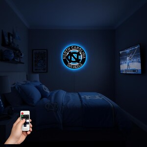May include: A round, black and blue LED wall light featuring the words "North Carolina Tar Heels" encircling the team logo. The light is mounted on a dark blue wall in a bedroom setting. A remote control is held in the foreground.