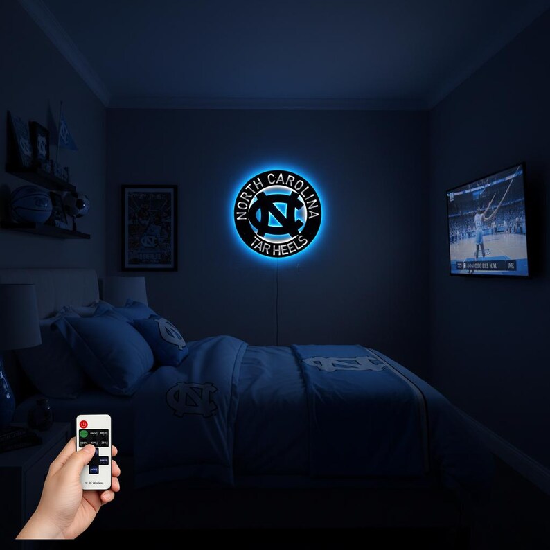 May include: A round, black and blue LED wall light featuring the words "North Carolina Tar Heels" encircling the team logo. The light is mounted on a dark blue wall in a bedroom setting. A remote control is held in the foreground.