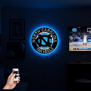 May include: A black and blue illuminated wall sign featuring the North Carolina Tar Heels logo. The sign is circular with the words "NORTH CAROLINA" and "TAR HEELS" around the logo. The logo is a stylized "NC" in white and blue.