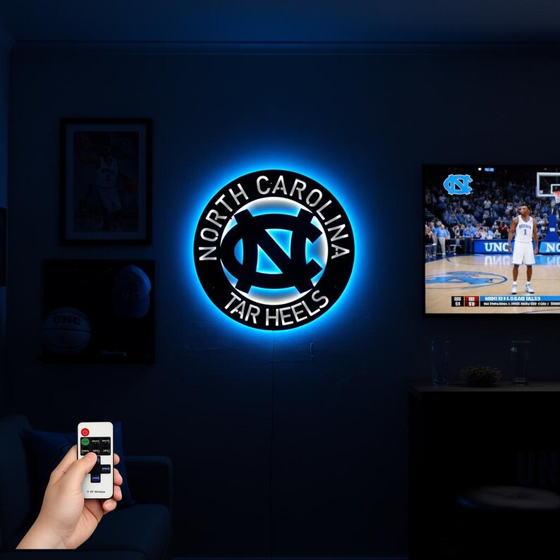 May include: A black and blue illuminated wall sign featuring the North Carolina Tar Heels logo. The sign is circular with the words "NORTH CAROLINA" and "TAR HEELS" around the logo. The logo is a stylized "NC" in white and blue.