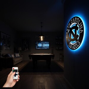 May include: A lit wall sign with the words "North Carolina Tar Heels" in a circular design. The sign is black with a blue neon glow around the edges. The sign is mounted on a dark gray wall in a room with a pool table and a TV.