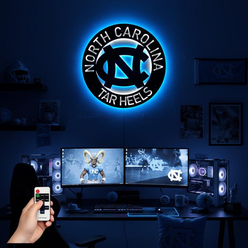 May include: A circular, black metal sign with the words "NORTH CAROLINA" and "TAR HEELS" encircling a blue and white logo. The sign is illuminated with a blue LED light, mounted on a dark blue wall. The image shows a gaming setup with two monitors.
