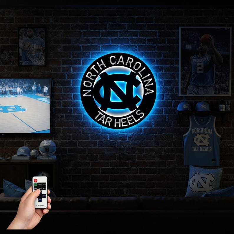 May include: A black metal wall art piece with the words "North Carolina Tar Heels" in a circular design. The logo is illuminated with a blue glow, mounted on a brick wall. Other Tar Heels merchandise is also visible.
