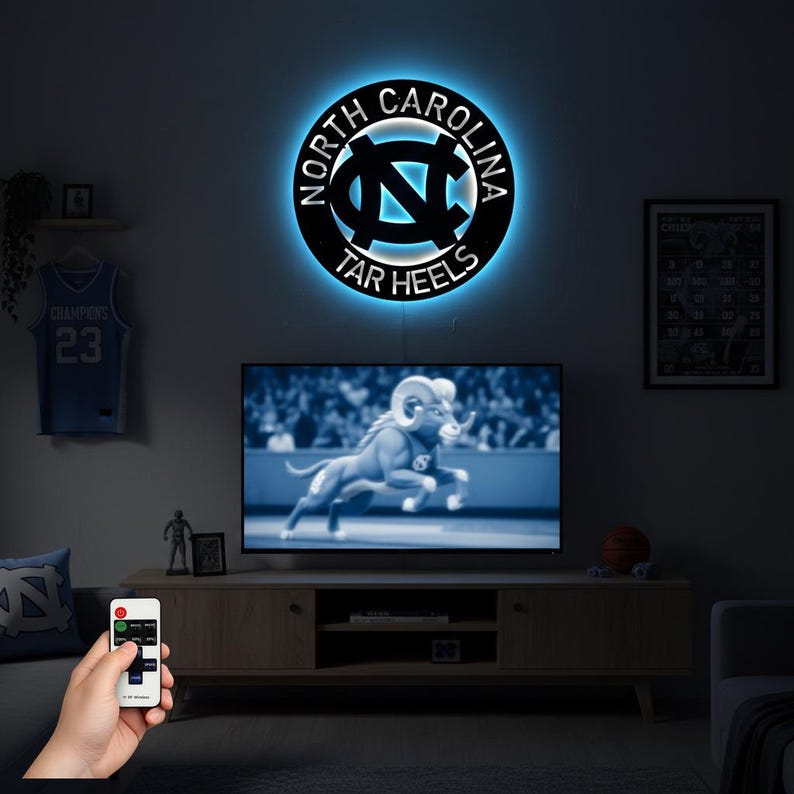 May include: A black metal wall art piece with the words "North Carolina Tar Heels" encircling the team logo, illuminated by a blue LED light. The logo is centered and also illuminated. A TV below displays a ram.