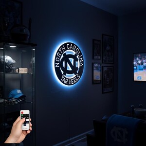 May include: A circular wall sign with the words "North Carolina Tar Heels" in white lettering. The sign has a blue neon backlight, and is mounted on a dark gray wall. A remote control is visible in the lower left corner.