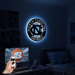May include: A black and blue illuminated wall sign featuring the words "North Carolina Tar Heels" in a circular design. The sign is mounted on a dark blue wall, with a remote control in the foreground.