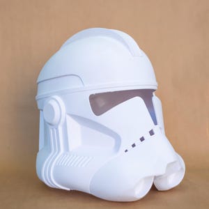 Puede incluir: Casco blanco de plástico moldeado, que recuerda a un casco de soldado de asalto. El casco presenta una visera, una parte superior redondeada y varios detalles moldeados. El diseño general es simétrico y limpio, centrado en la forma icónica.