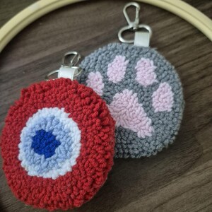 May include: Two handmade keyrings. One is red with a blue and white eye design. The other is grey with a pink paw print. Both have silver-coloured key rings and white fabric loops.
