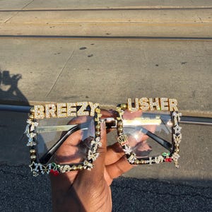Puede incluir: Gafas con lentes transparentes con las palabras "BREEZY" y "USHER" en letras doradas y brillantes. Las monturas son negras con adornos dorados y brillantes, incluyendo estrellas, mariposas y cerezas.