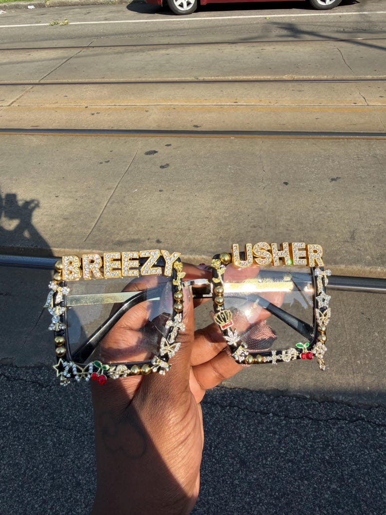 Puede incluir: Gafas con lentes transparentes con las palabras "BREEZY" y "USHER" en letras doradas y brillantes. Las monturas son negras con adornos dorados y brillantes, incluyendo estrellas, mariposas y cerezas.