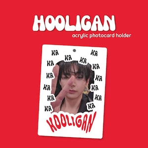 May include: A rectangular white acrylic photocard holder featuring the text HOOLIGAN in red at the bottom and repeated black KA text patterns. The center displays a photograph of a person making a peace sign. The top has a small circular hole.