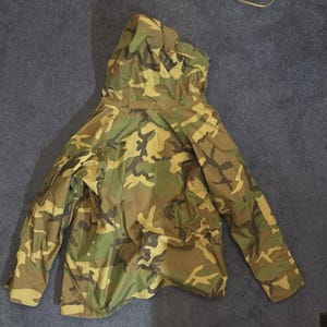 May include: A camouflage pattern jacket with a hood, in shades of green, brown, and tan. The jacket is lying on a dark blue surface. A straw hat with a brown strap is visible in the upper right corner.