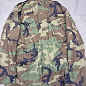 May include: Camouflage pattern jacket with multiple pockets and a high collar. The jacket features a mix of green, brown, and black shapes. The jacket has a button and zipper closure.