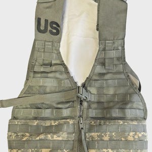 May include: A military-style tactical vest in shades of green and tan camouflage. The vest features a front zipper, multiple straps, and the letters "US" printed in black on the shoulder area. The vest is designed with a modular webbing system.