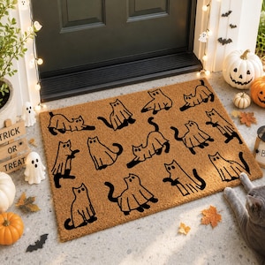 May include: A rectangular tan coir doormat featuring a repeating black line art pattern of cats wearing ghost costumes. The cats are depicted in various poses, including sitting, stretching, and standing, all draped in white-style ghost sheets.