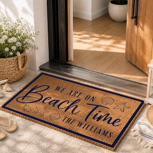 May include: A rectangular tan coir doormat with a dark blue border. The mat features the text "WE ARE ON Beach Time" and "THE WILLIAMS" in dark blue script and block lettering, accompanied by illustrations of seashells and starfish.