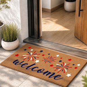 May include: A rectangular tan coir doormat featuring the word welcome in dark blue script. The design includes red and blue firework bursts and small red, white, and blue stars scattered across the surface, creating a festive patriotic aesthetic.