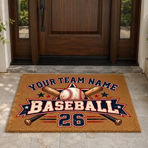 May include: A rectangular tan coir doormat featuring a baseball graphic with two crossed wooden bats, a red and blue banner with the text BASEBALL, the phrase YOUR TEAM NAME, and the number 26 in a bold, dark blue font with red outlines.