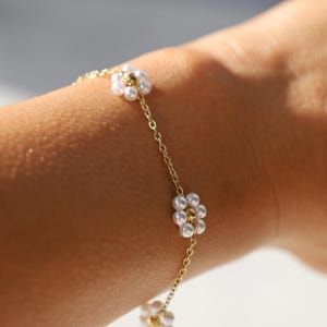 May include: A gold-toned chain bracelet featuring multiple flower-shaped charms. Each flower is constructed from small, round, white faux pearls arranged in a circular pattern around a central gold-toned bead. The delicate chain links connect each floral element.