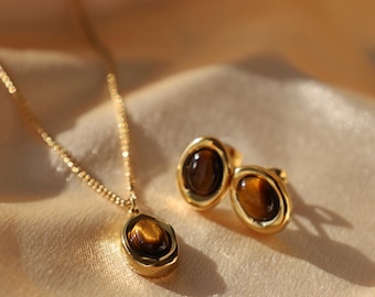 May include: A gold-toned necklace featuring an oval pendant with a brown tiger's eye gemstone and a matching pair of stud earrings, each containing a single oval tiger's eye stone set within a smooth, polished gold-colored metal frame.