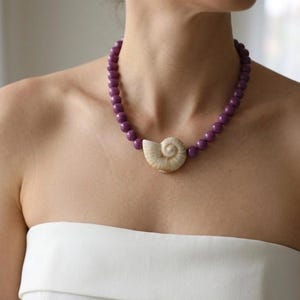 May include: A necklace featuring a series of smooth, round, purple beads strung together. The centerpiece of the necklace is a single, natural, cream-colored spiral seashell positioned at the front, creating a distinct contrast with the purple beads.