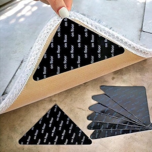 May include: Black triangular rug grippers with the text "on floor" repeated across the surface. The grippers are shown in use, holding the corner of a rug. A stack of additional grippers is also visible.