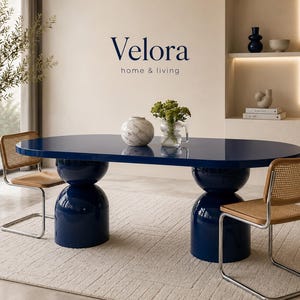 Puede incluir: Una mesa de comedor ovalada de color azul marino brillante con dos grandes bases de pedestal esféricas apiladas. Dos sillas con marcos cromados y respaldos y asientos de rejilla tejida de color marrón claro se sitúan a ambos lados. El texto Velora home & living es visible.