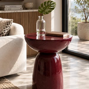 Puede incluir: Una mesa auxiliar de color rojo oscuro brillante con una parte superior redondeada en forma de cuenco y una base cilíndrica cónica. La mesa presenta un acabado liso y reflectante con una forma sólida y escultural sobre el suelo.