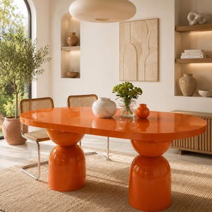 Puede incluir: Una mesa de comedor ovalada con un acabado naranja brillante, sostenida por dos bases de pedestal redondeadas y apiladas. La superficie de la mesa es lisa y reflectante, con un jarrón de mármol blanco, un jarrón de cristal con flores y un pequeño jarrón naranja.
