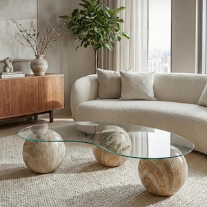 Op de afbeelding: Een moderne woonkamer met een glazen salontafel ondersteund door drie bolvormige bases met een marmerachtig patroon. Een crèmekleurige bank met kussens en een houten kast met een vaas en decoratieve items zijn ook zichtbaar. De tafel is ongeveer 100 cm lang.
