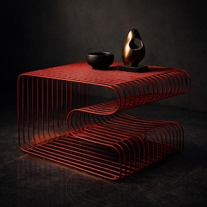 Peut inclure: Une table d'appoint moderne en métal rouge avec un design incurvé unique. La table présente une structure à deux niveaux faite de fines barres parallèles. Un bol noir et une sculpture dorée sont posés dessus. La table mesure environ 61 cm de long.