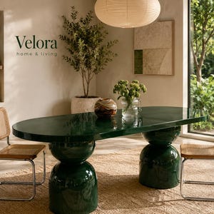 Pode incluir: Uma mesa de jantar oval verde-escura brilhante com duas grandes bases arredondadas, acompanhada por duas cadeiras com estruturas de madeira clara, assentos e encostos de palhinha trançada, com o texto Velora home & living na parede.