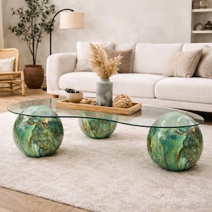 May include: A coffee table featuring a clear, oval-shaped glass top supported by three large, spherical bases with a marbled green and gold finish. The table is positioned on a light-colored rug, showcasing a smooth, reflective surface and rounded forms.