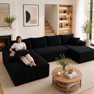 Puede incluir: Un gran sof&aacute; modular de esquina de pana negro con varios cojines. El sof&aacute; est&aacute; dispuesto en forma de L, con una persona sentada en la secci&oacute;n de la esquina. Una mesa de centro redonda de madera con libros y un jarr&oacute;n con flores est&aacute; frente al sof&aacute;.