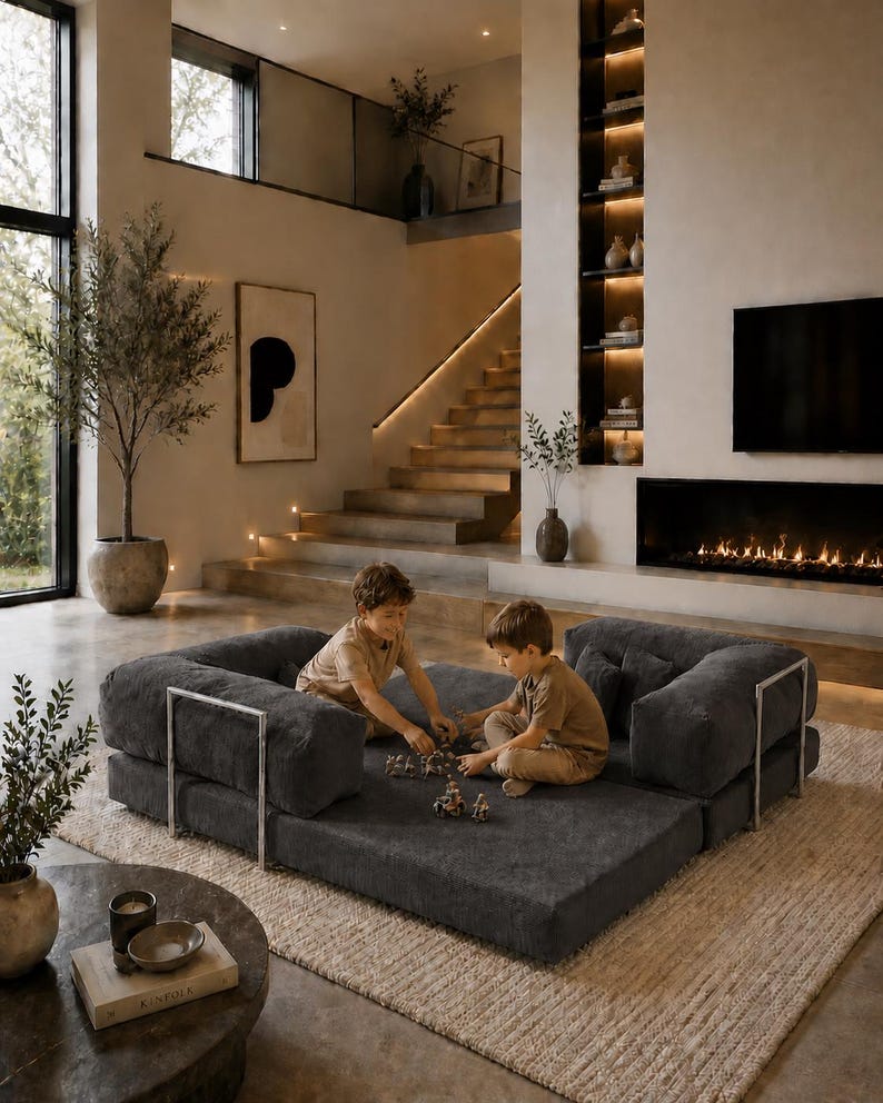 Puede incluir: Un sal&oacute;n moderno con un gran sof&aacute; modular gris oscuro. Dos ni&ntilde;os juegan en el sof&aacute;. La habitaci&oacute;n presenta una paleta de colores neutros, una chimenea y una escalera que conduce a un segundo nivel.