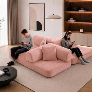 Puede incluir: Sal&oacute;n moderno con un sof&aacute; modular rosa y una mesa de centro negra. Una persona est&aacute; sentada en el sof&aacute;, usando un tel&eacute;fono inteligente. La habitaci&oacute;n incluye una gran obra de arte abstracta y una l&aacute;mpara colgante. El sof&aacute; tiene varios cojines.
