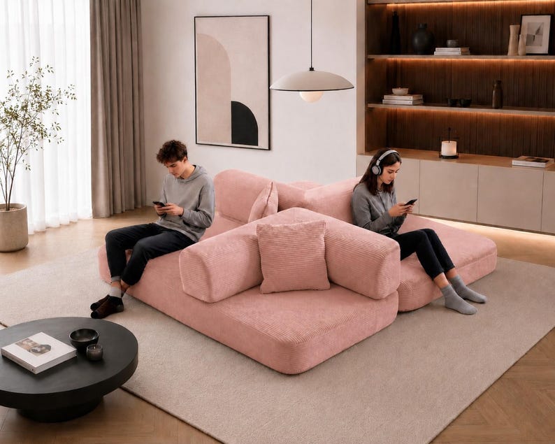 Puede incluir: Sal&oacute;n moderno con un sof&aacute; modular rosa y una mesa de centro negra. Una persona est&aacute; sentada en el sof&aacute;, usando un tel&eacute;fono inteligente. La habitaci&oacute;n incluye una gran obra de arte abstracta y una l&aacute;mpara colgante. El sof&aacute; tiene varios cojines.