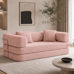 Puede incluir: Un sof&aacute; de pana rosa con dos cojines a juego. El sof&aacute; tiene un dise&ntilde;o moderno con un reposabrazos de metal y est&aacute; colocado sobre una alfombra de color claro. Una mesa de centro redonda y objetos decorativos est&aacute;n en el fondo.