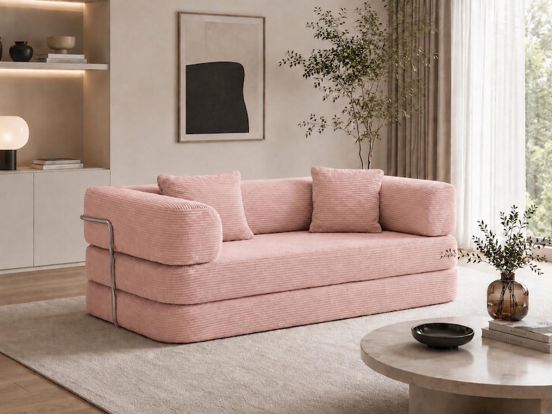 Puede incluir: Un sof&aacute; de pana rosa con dos cojines a juego. El sof&aacute; tiene un dise&ntilde;o moderno con un reposabrazos de metal y est&aacute; colocado sobre una alfombra de color claro. Una mesa de centro redonda y objetos decorativos est&aacute;n en el fondo.