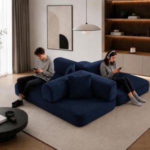 May include: A modern living room featuring a large, modular navy blue sofa with multiple cushions. A round black coffee table sits in front of the sofa. A person is sitting on the sofa using a smartphone. A large abstract painting hangs on the wall.