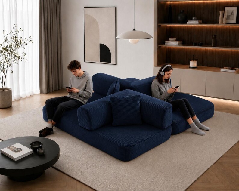 May include: A modern living room featuring a large, modular navy blue sofa with multiple cushions. A round black coffee table sits in front of the sofa. A person is sitting on the sofa using a smartphone. A large abstract painting hangs on the wall.