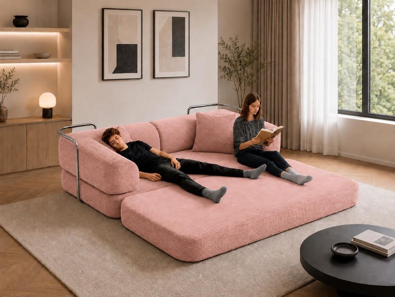 Puede incluir: Un gran sof&aacute; modular rosa de pana con una extensi&oacute;n de chaise longue. El sof&aacute; tiene un dise&ntilde;o moderno con reposabrazos de metal. Una persona est&aacute; acostada en la chaise longue y otra est&aacute; leyendo un libro. La habitaci&oacute;n tiene una paleta de colores neutros.
