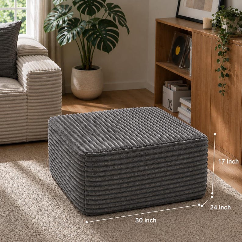 Puede incluir: Un otom&aacute;n de pana gris, de 76 cm de largo, 61 cm de ancho y 43 cm de alto. El reposapi&eacute;s rectangular est&aacute; sobre una alfombra beige. Un sof&aacute; y un mueble de madera est&aacute;n en el fondo, con una gran planta en maceta.