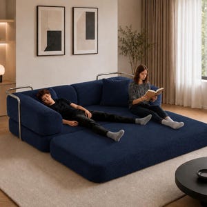 May include: A modern, modular navy blue sofa with a chaise lounge extension. The sofa features a textured fabric and chrome accents. Two people are relaxing on the sofa, one reading a book and the other resting. The sofa sits on a light-colored rug.