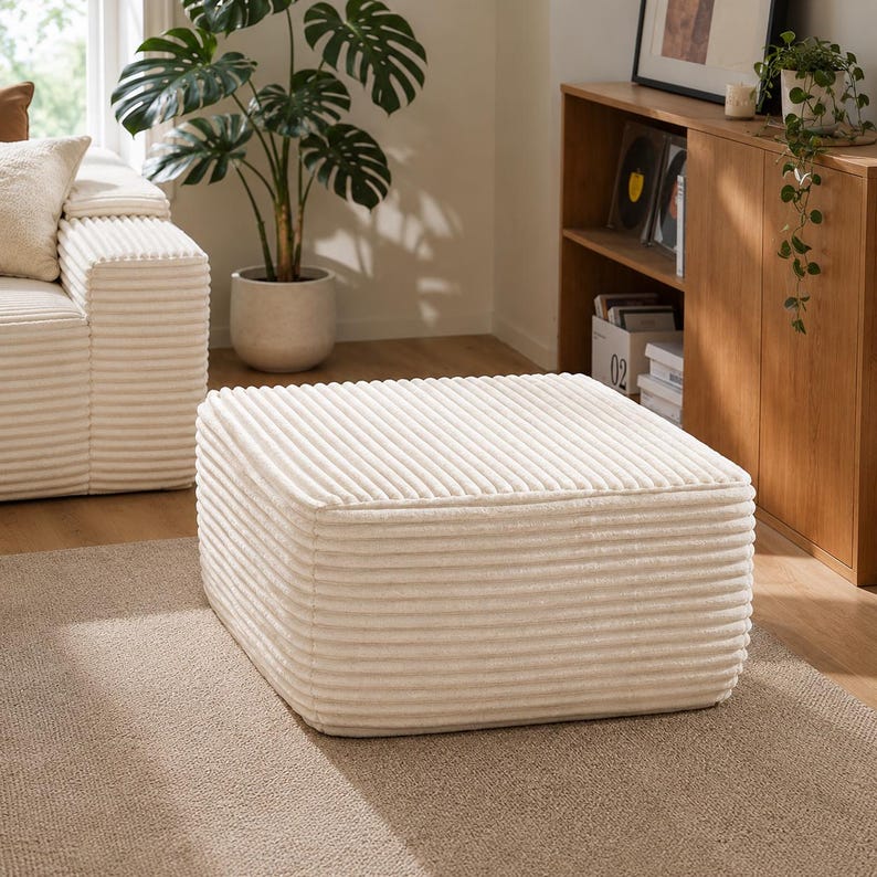 Puede incluir: Un otom&aacute;n de pana color crema, de forma cuadrada. Presenta una textura acanalada y est&aacute; colocado sobre una alfombra de tonos neutros. El dise&ntilde;o es simple y moderno, adecuado para la decoraci&oacute;n del sal&oacute;n. Dimensiones: aprox. 70 cm x 70 cm.