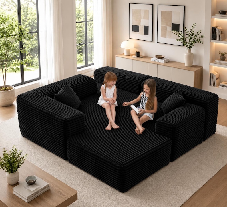 Puede incluir: Un gran sof&aacute; modular de pana negro con m&uacute;ltiples secciones y cojines. El sof&aacute; est&aacute; dispuesto en forma de L y se coloca sobre una alfombra de color claro. Dos ni&ntilde;os peque&ntilde;os est&aacute;n sentados en el sof&aacute;.