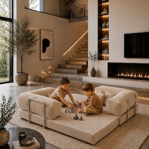 Puede incluir: Un sal&oacute;n moderno con un sof&aacute; modular beige, dos ni&ntilde;os jugando y una paleta de colores neutros. La habitaci&oacute;n cuenta con una chimenea, una ventana grande y una escalera que conduce a un segundo nivel. Una mesa de centro redonda con un libro y velas est&aacute; en primer plano.